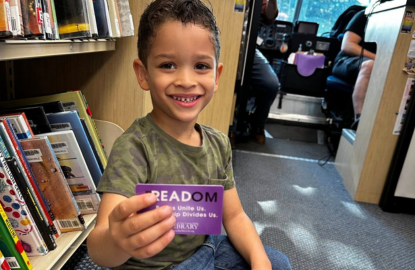 A young child holding up a library card while smiling